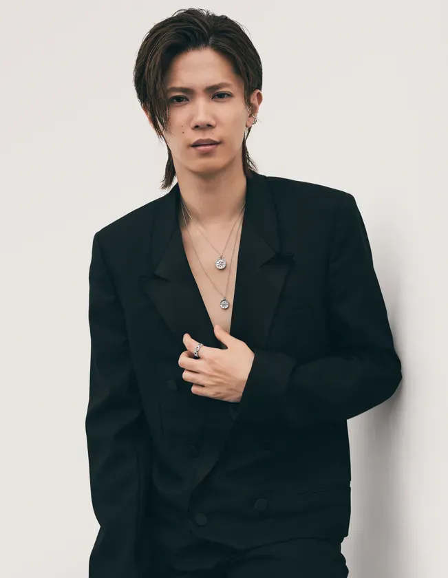 黒いジャケットを着た男性が白い壁に寄りかかって立っている写真です。男性は、長めの髪型で、ネックレスと指輪を着用しています。