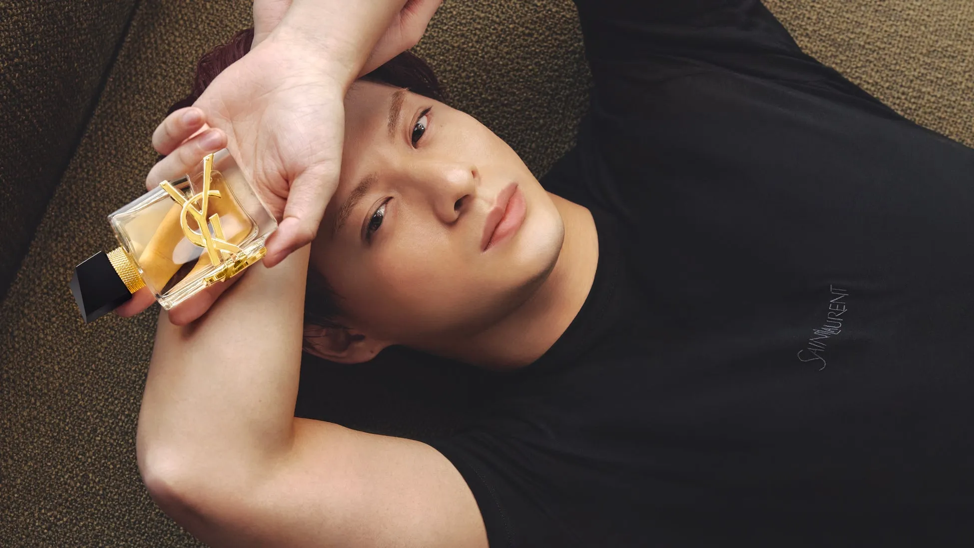 男性が香水を持ち、横たわっている写真。彼は黒い服を着ており、カメラを見つめている。香水は彼の手に持たれており、彼の顔の近くに配置されている。