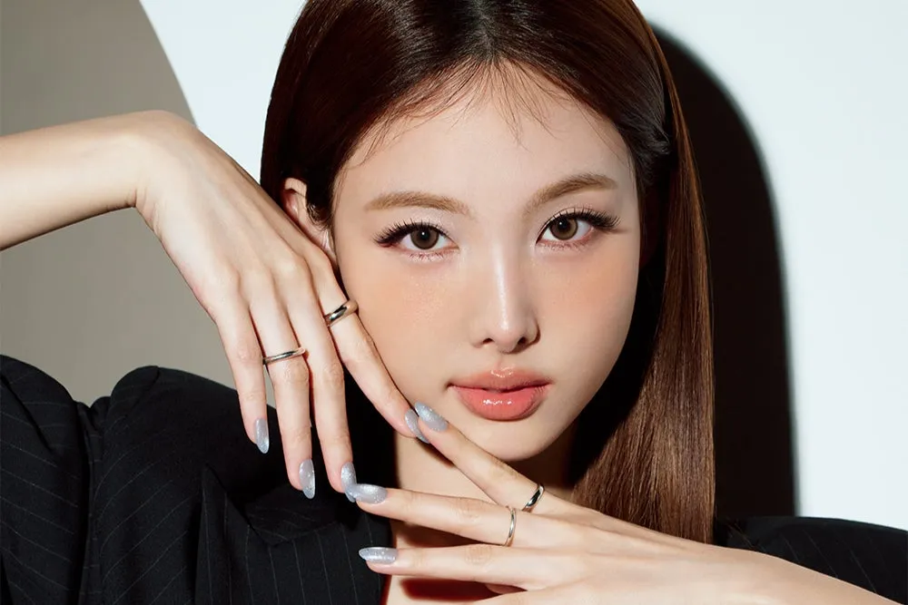 女性のポートレート。長い茶色の髪で、顔に手を添えている。彼女はメイクをしており、唇は光沢がある。指には指輪がはめられている。