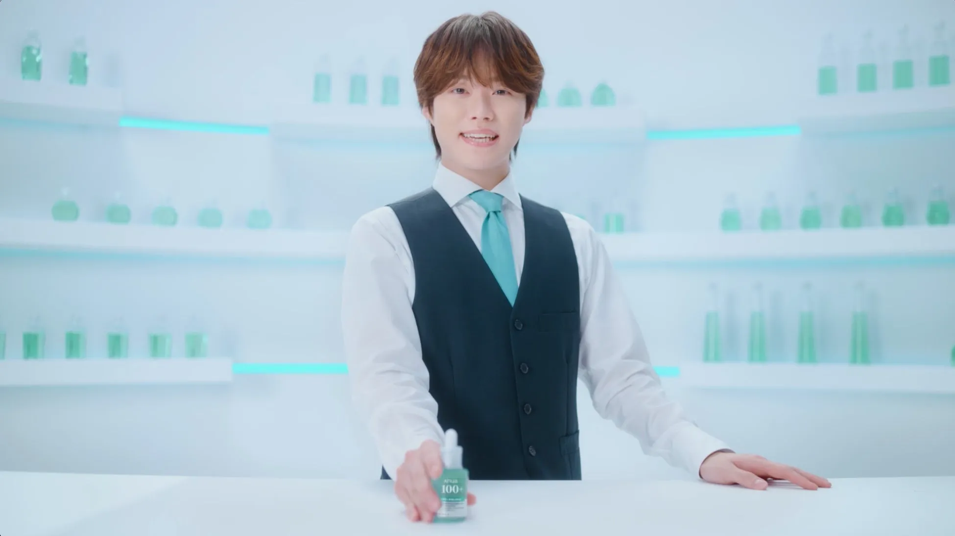 若い男性が美容液のボトルを持ち、笑顔でカメラを見ている様子。背景には、棚に並んだ緑色の液体が入ったボトルが映っている。