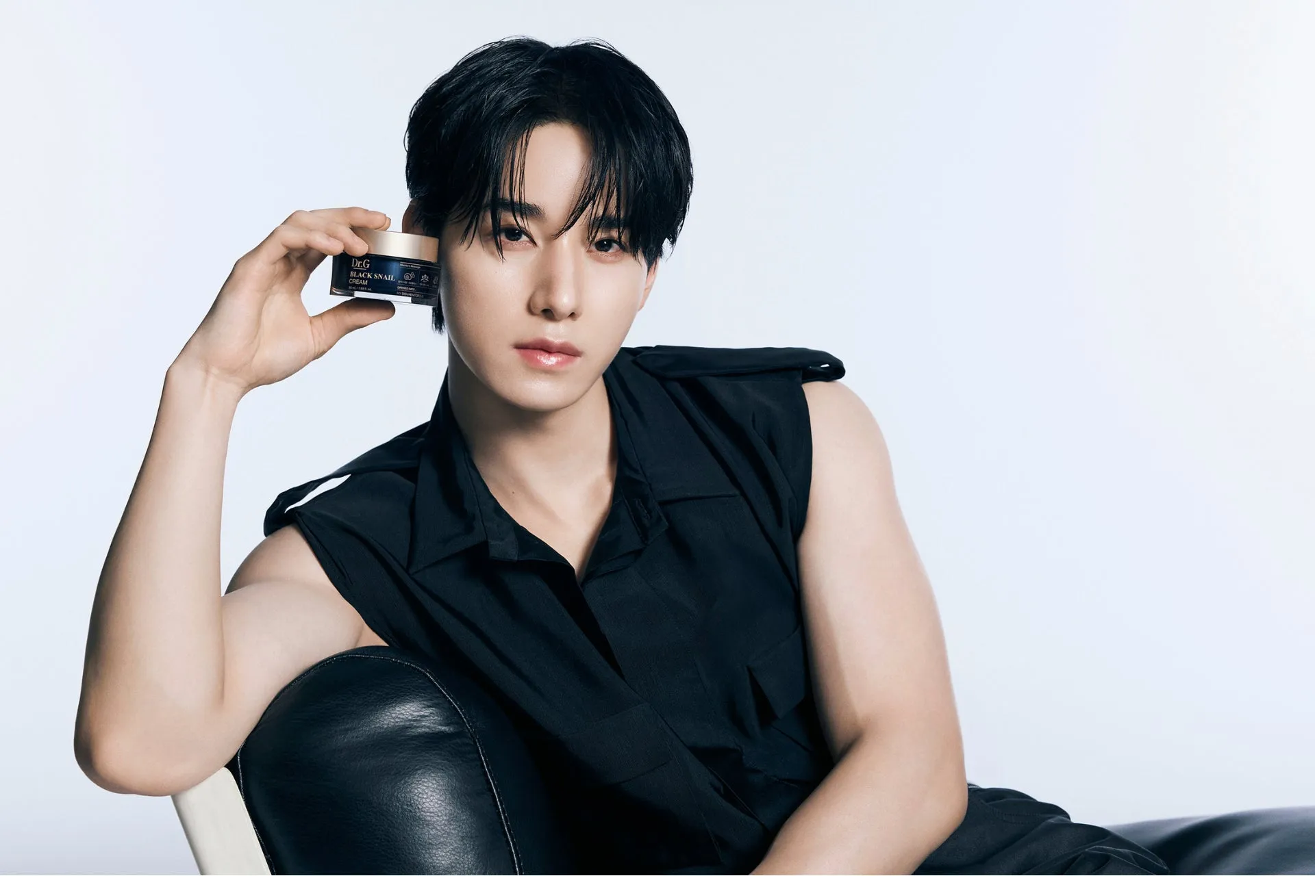 黒いノースリーブのシャツを着た男性が、顔の近くに化粧品の容器を持っています。彼は革張りの椅子に座っています。