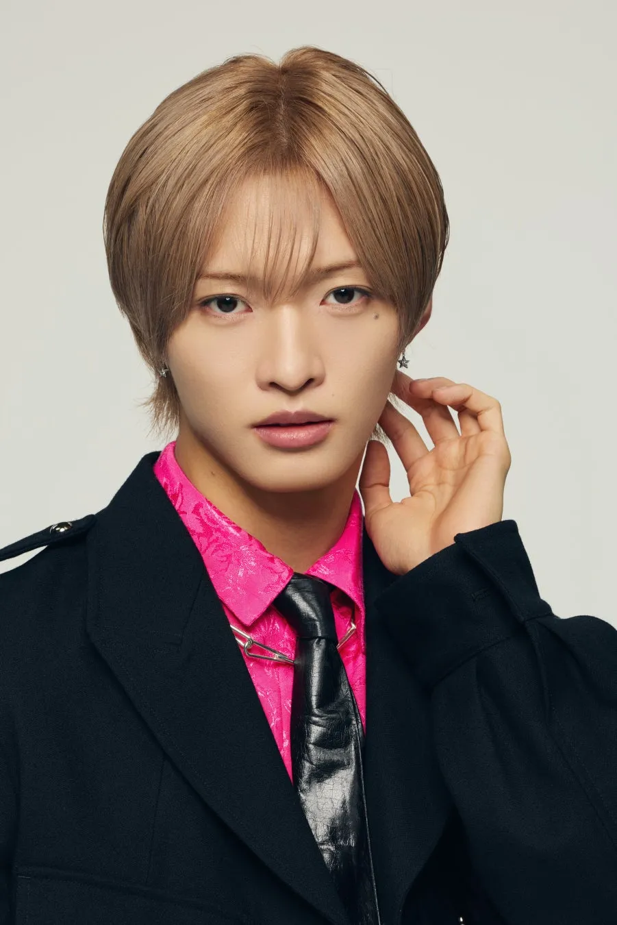 男性が正面を向いてポーズをとっているポートレート写真。彼はピンクのシャツと黒いネクタイを着ており、黒いジャケットを着ています。彼の髪は短く、ブロンドです。背景は白。