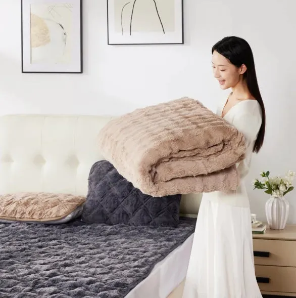 女性が毛布と枕を持っている画像です。ベッドの上にはグレーの毛布と枕が置かれています。壁には絵が飾られています。