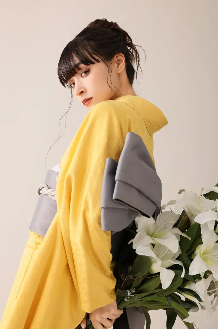 黄色い着物を着た女性が、白いユリの花束を持ってポーズをとっている。女性はカメラ目線で、髪はアップにまとめられ、化粧をしている。