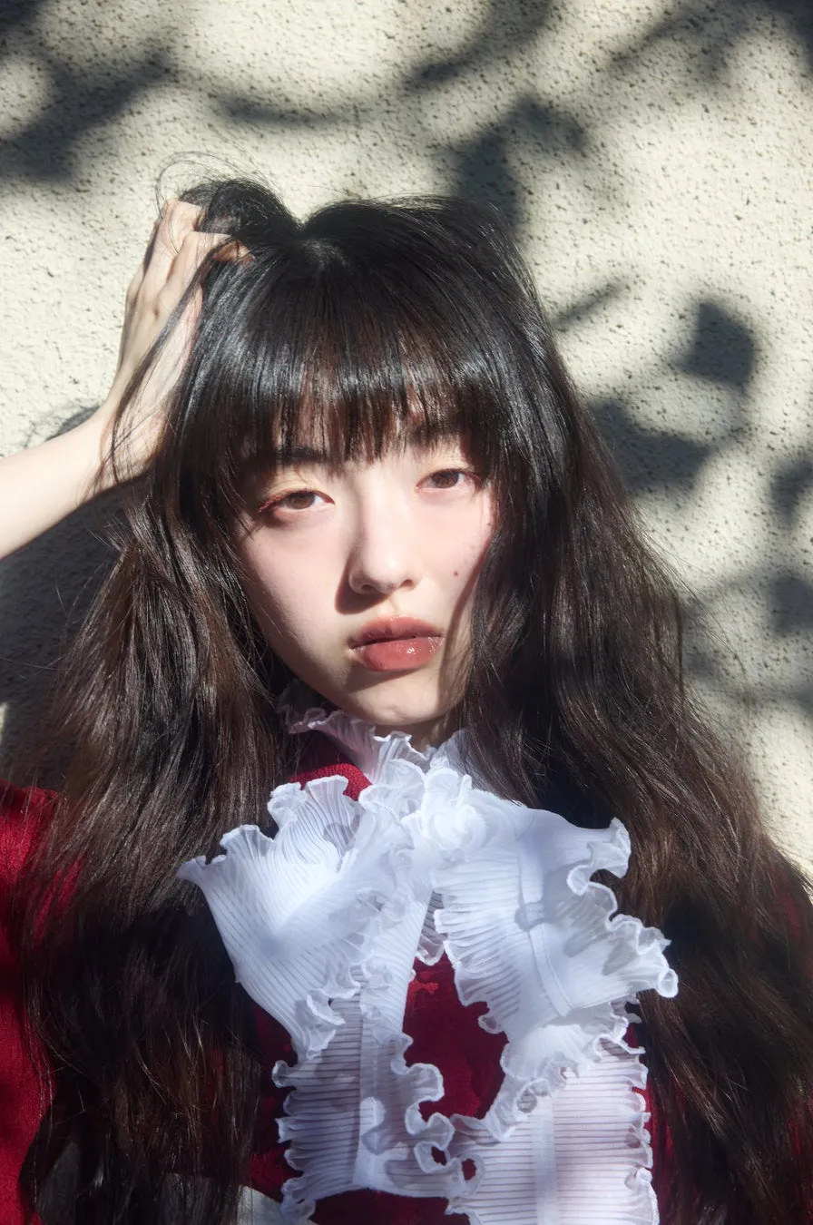 女性のポートレート。長い黒髪で、前髪があり、手で髪に触れている。白いフリルの襟と赤い服を着ている。背景は日差しによる影がある壁。