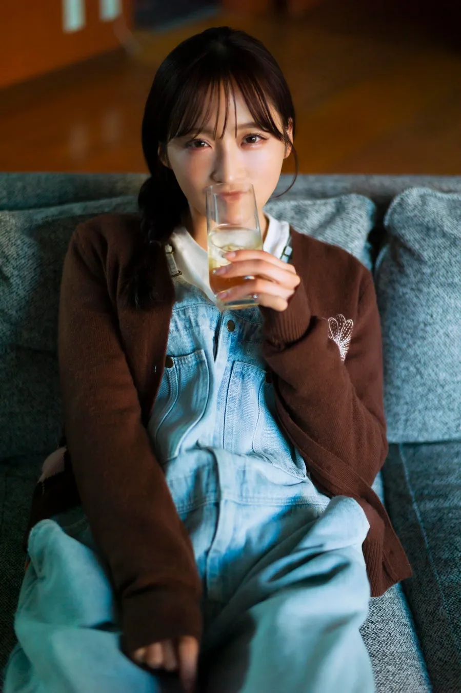 女性がグラスに入った飲み物を飲んでいる写真です。女性はデニムのオーバーオールと茶色のカーディガンを着ており、ソファに座っています。