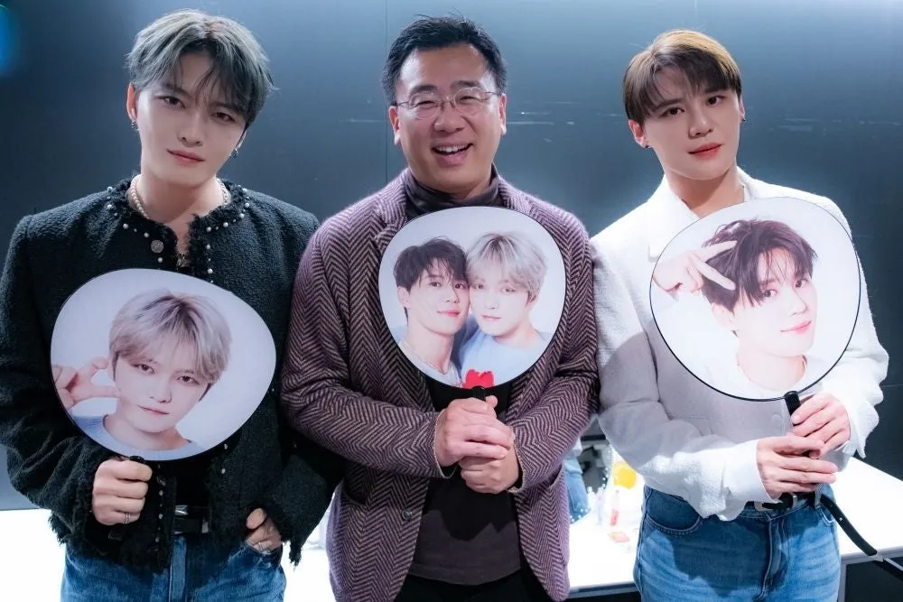 3人の人物がうちわを持って立っている写真です。うちわには、それぞれ異なる男性の写真が印刷されています。真ん中の男性は眼鏡をかけています。