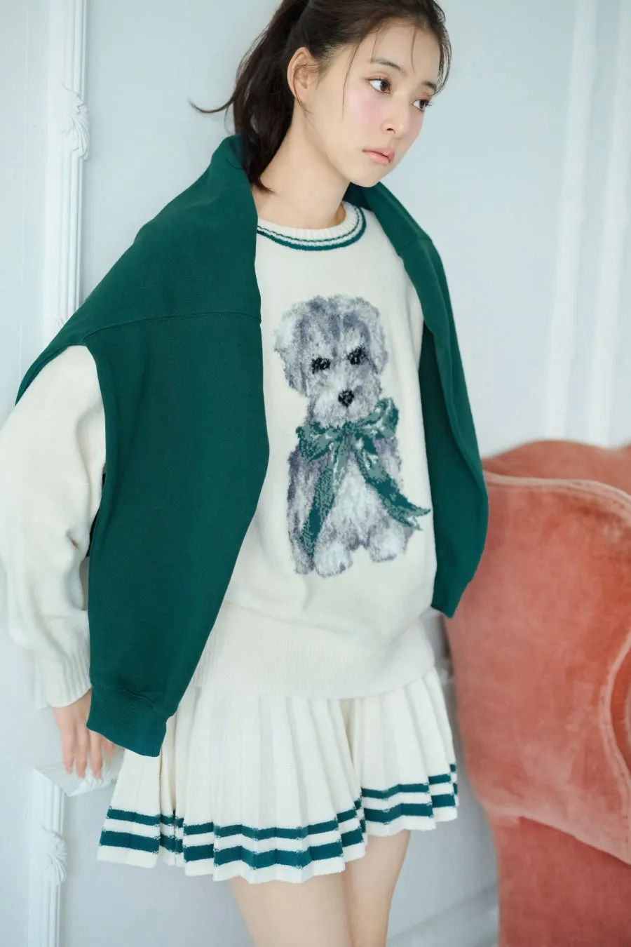 犬の絵が描かれたセーターとプリーツスカートを着た女性が、グリーンのジャケットを肩にかけて立っている様子を捉えた写真です。背景には白い壁とピンクの椅子が見えます。