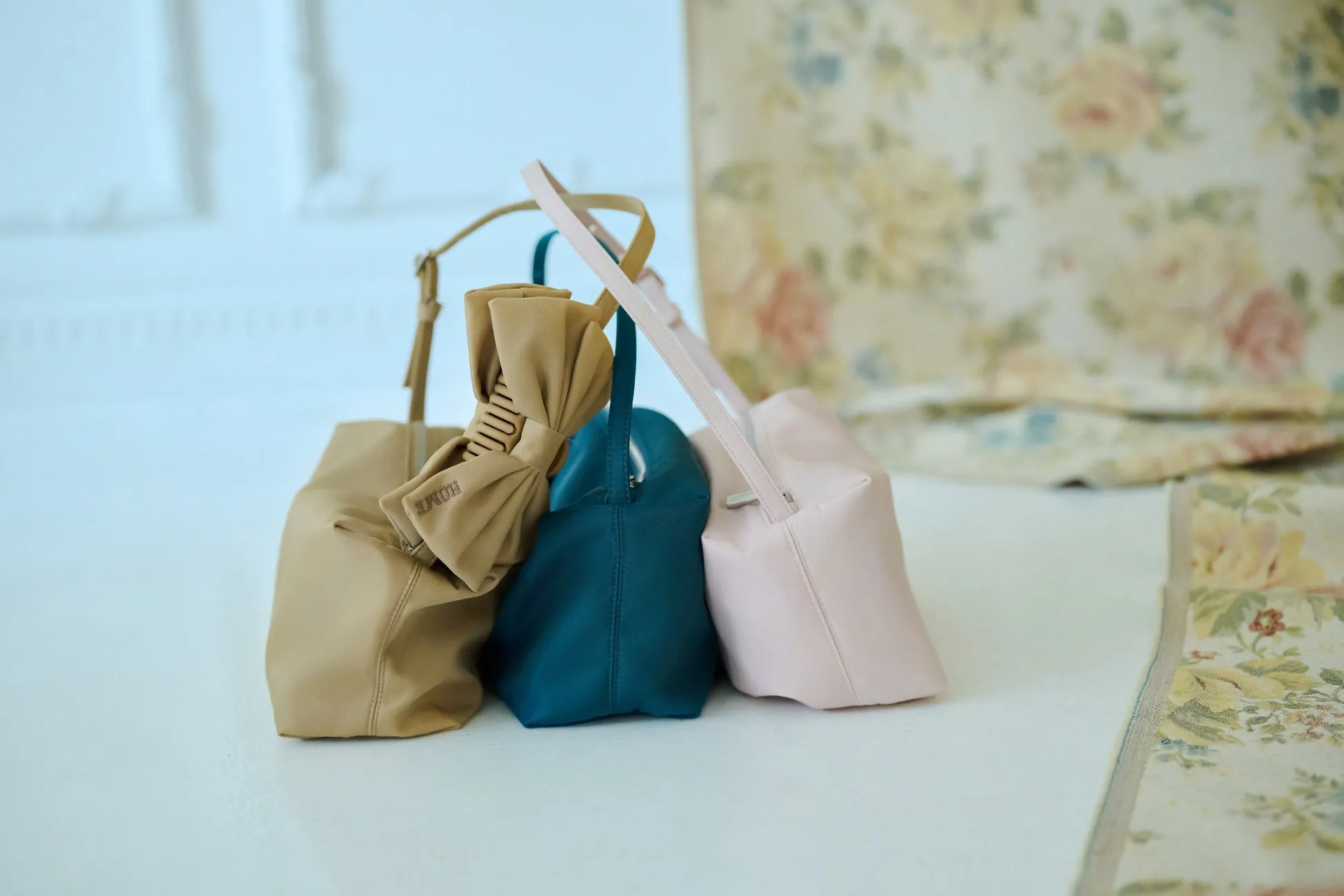 3つのカラフルなバッグが並んだ写真。左のバッグはベージュで、リボンのような装飾と「HOME」の文字が特徴。中央は濃い青色、右は薄いピンク色。背景には花柄のカーテンが見える。