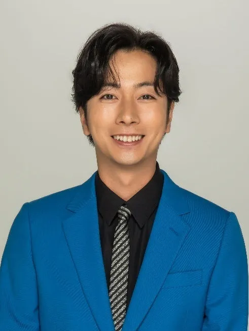 青いジャケットを着た男性のポートレートです。彼は黒いシャツとストライプのネクタイを着ており、笑顔でカメラを見ています。