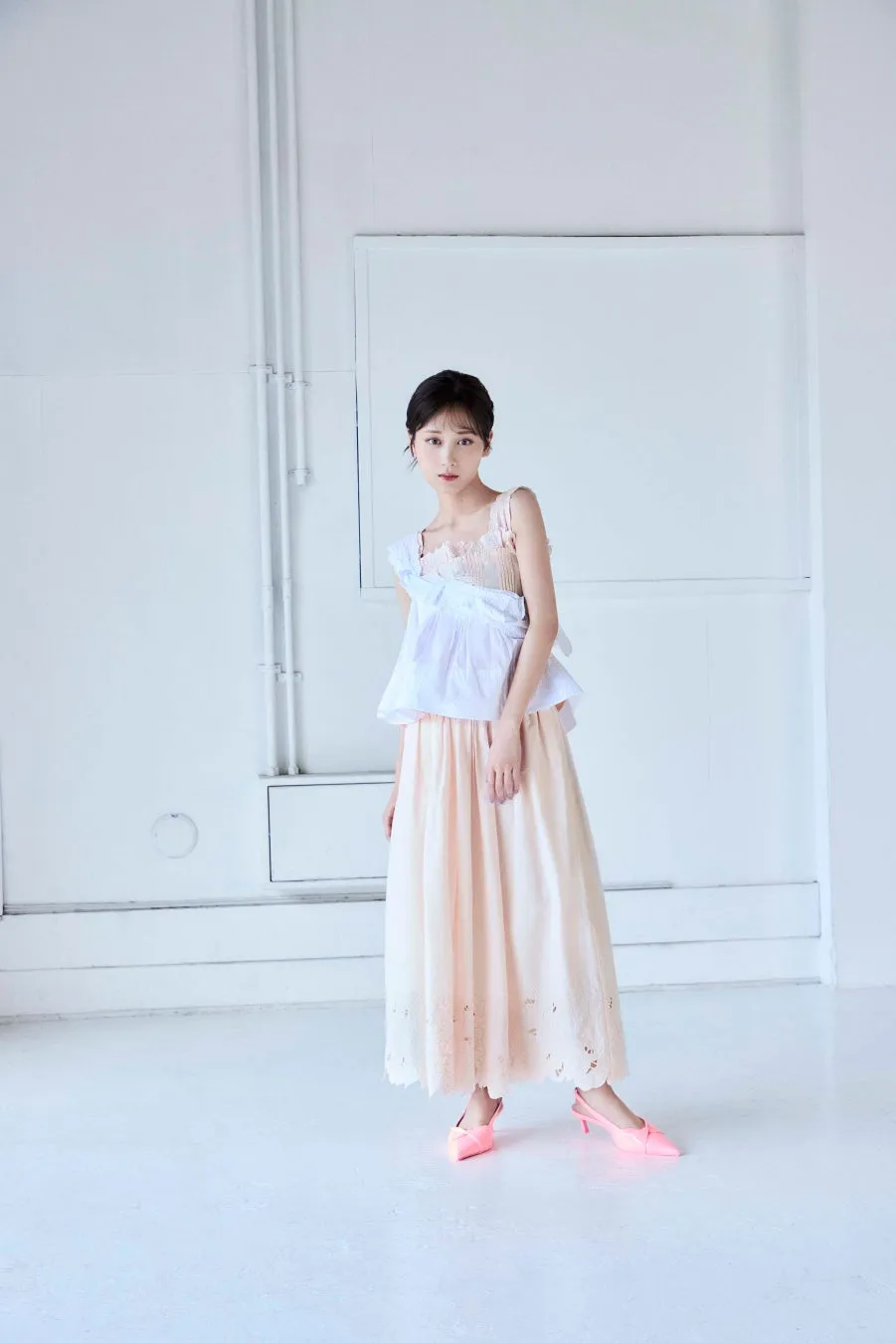 白い壁を背景に、ピンクのスカートと白いトップスを着た女性が立っている写真です。
