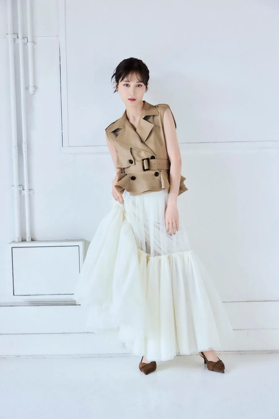 白い壁を背景に、女性がベージュのノースリーブジャケットと白いチュールのスカートを着用しています。