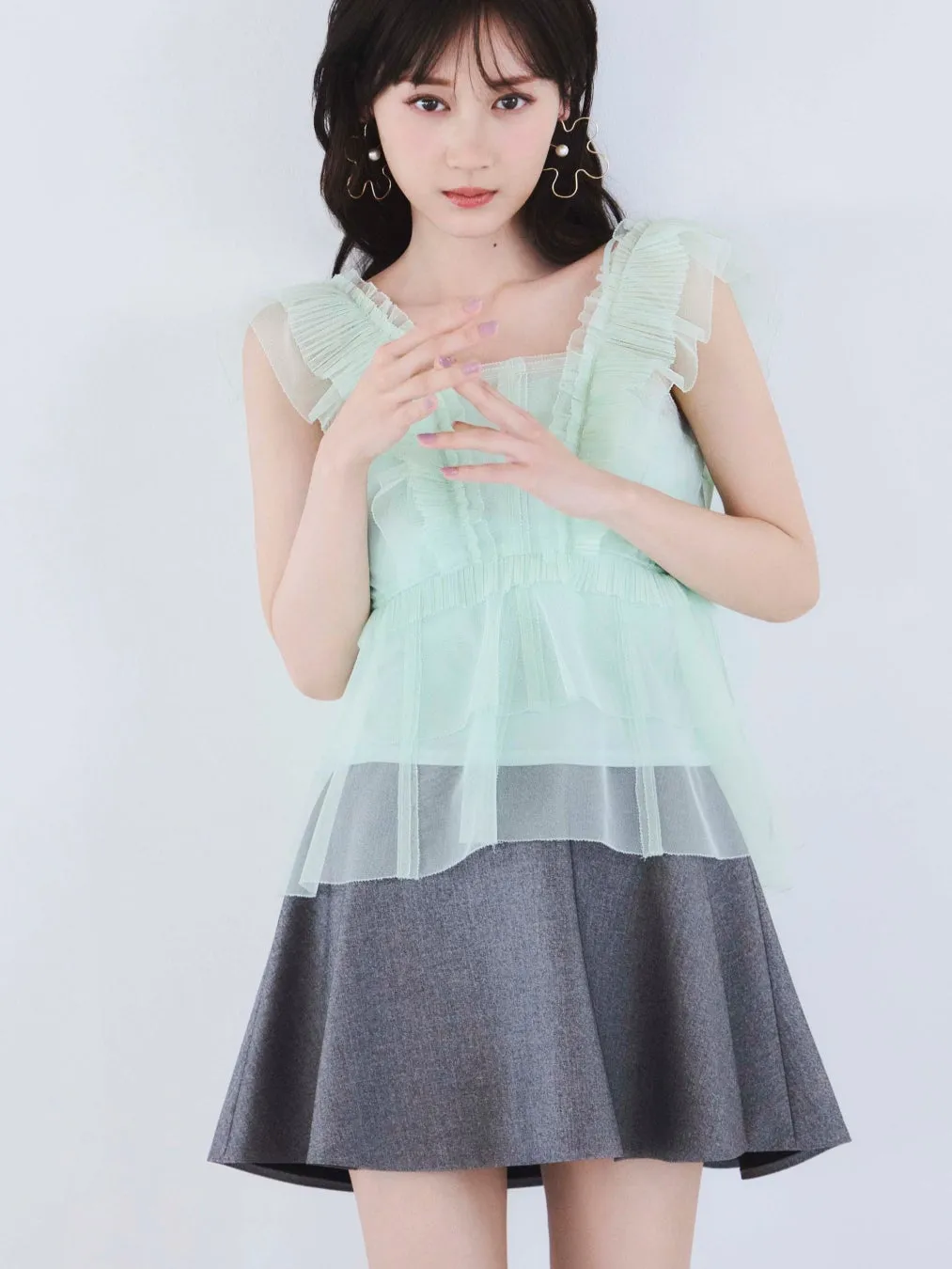 女性が白い壁の前に立っており、ミントグリーンのトップとグレーのスカートを着ています。
